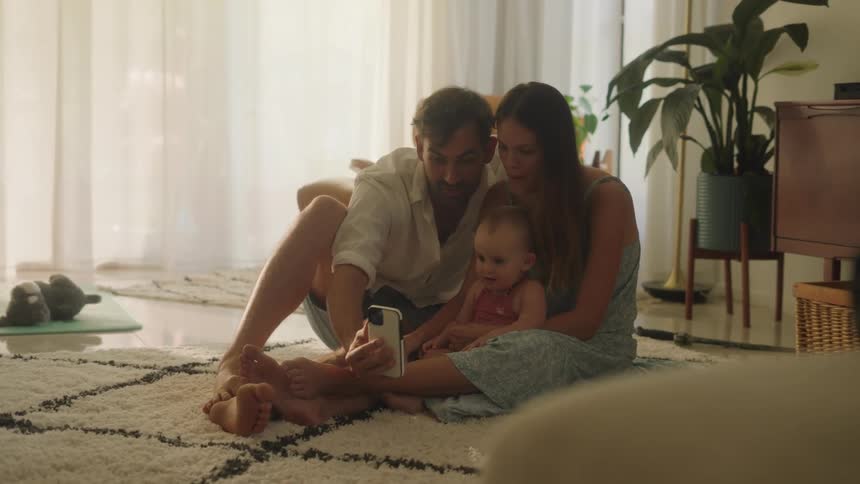 Father with white shirt and shorts take selfie with smartphone with mother in light blue dress and daughter sitting on the rug in the living room at home