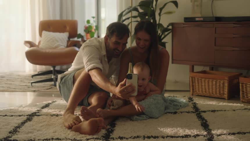 Young parents with their baby girl sitting on the floor laughingand taking a selfie of the family