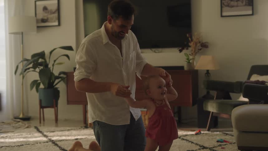 A father on his knees walking his baby daughter with his hands on carpet in the living room