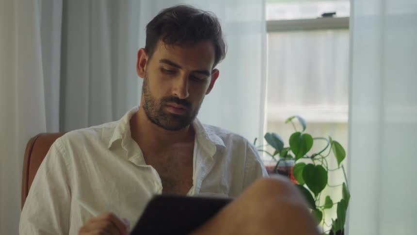 Man with stubble beard and white shirt sits in armchair using tablet in room