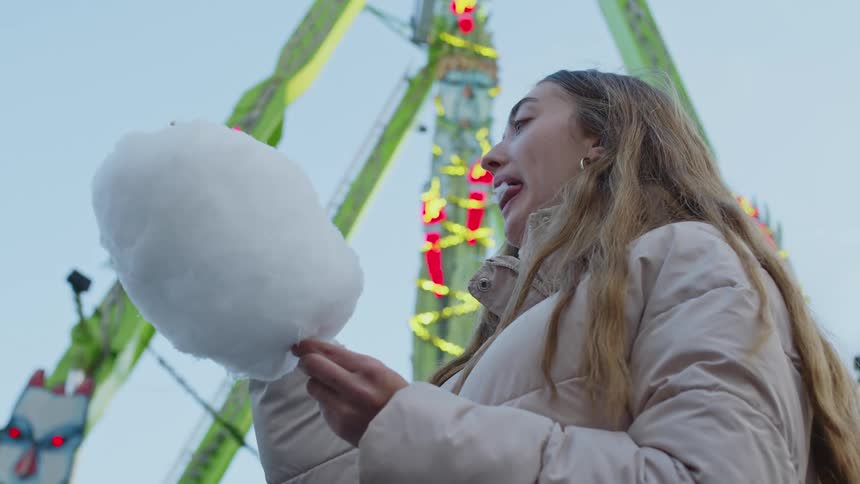 A woman eats cotton candy while standing by a carnival ride
