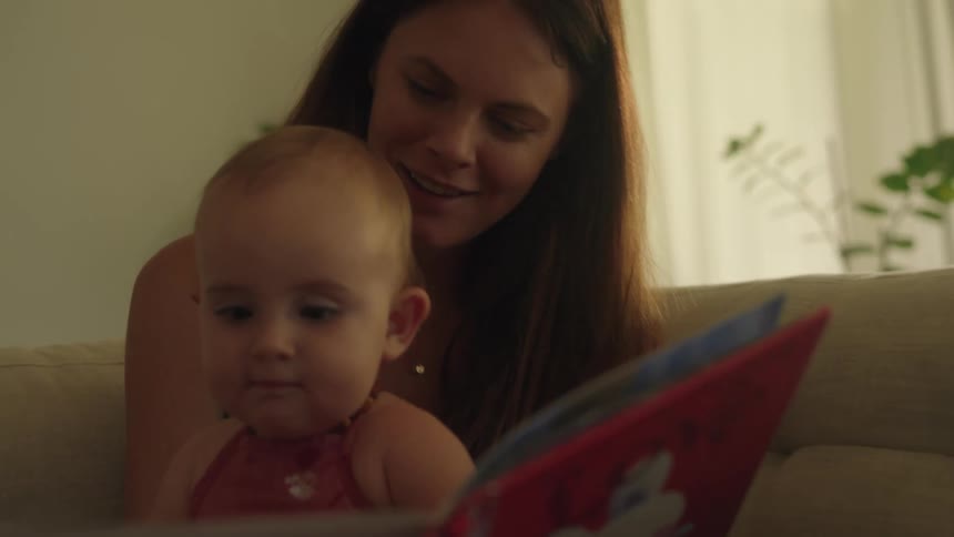 Woman is sitting on the living room couch with her baby looking at a book