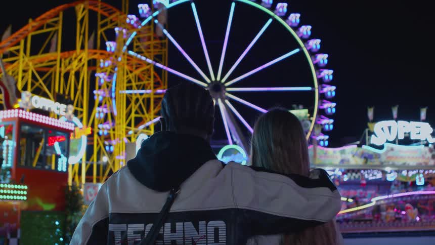 Man stands with arm around his date as they watch amusement park rides at night
