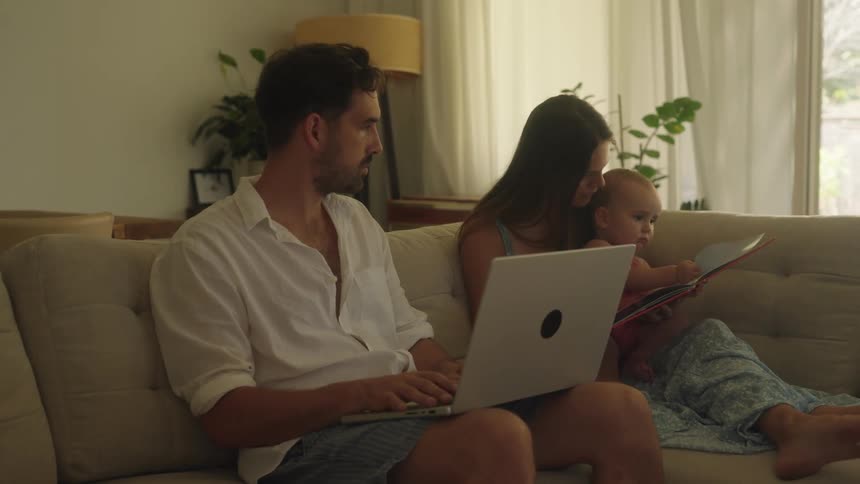 A woman reads a book to her baby while her husband uses a laptop beside them
