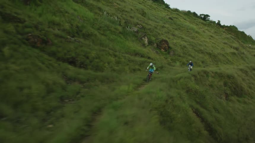 Two people on bicycles ride along the cliff of an overgrown green mountain