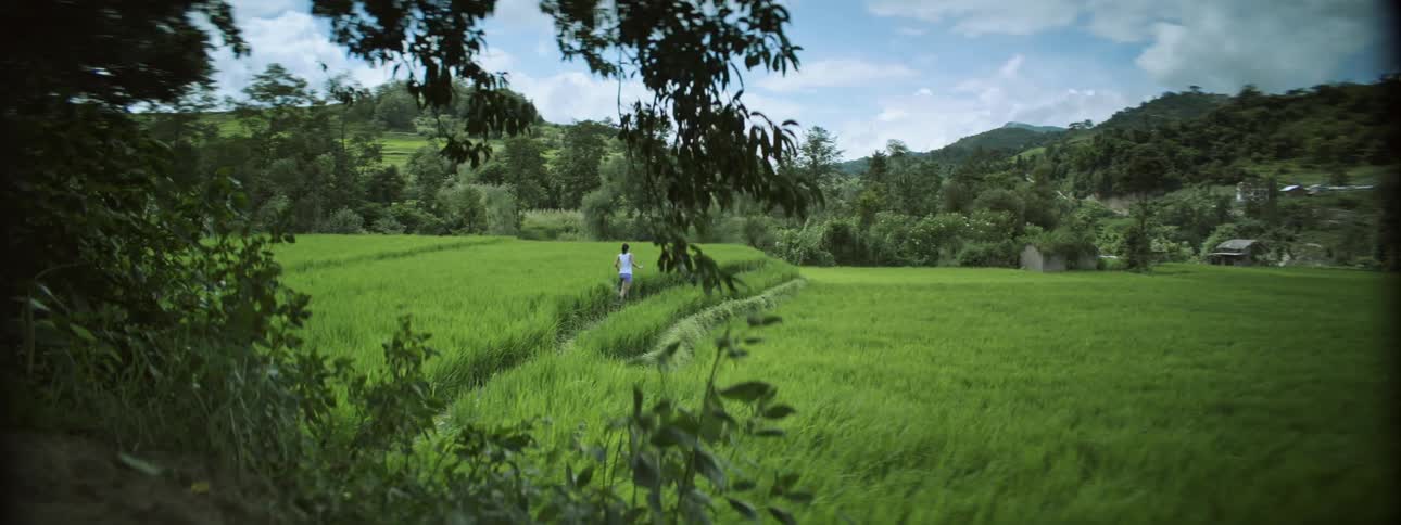 A woman jogs on a path through a grassy field toward a distant village and forest