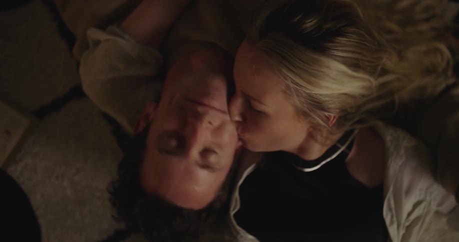 Blonde woman with black top and necklace and man lie on the floor and kiss each other