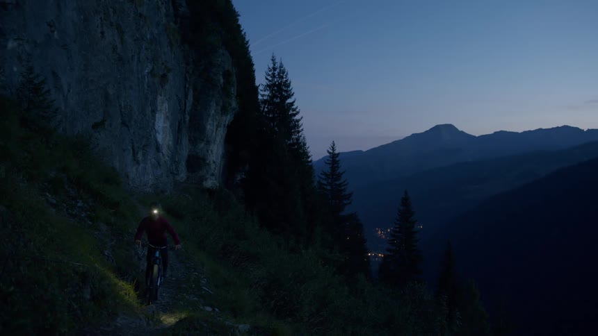 Cyclist gets off mountain bike on slope at base of cliff overlooking foggy town at night and starts guiding bike