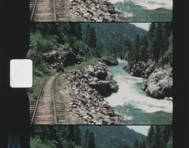 A train track with a cart on it next to a rapidly flowing river flowing through mountains