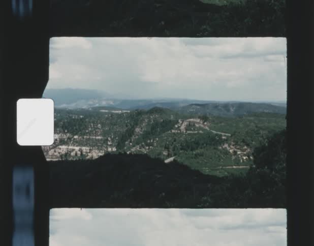 Looking over a ledge at forested mountains under a cloudy sky and there are mountains on the horizon