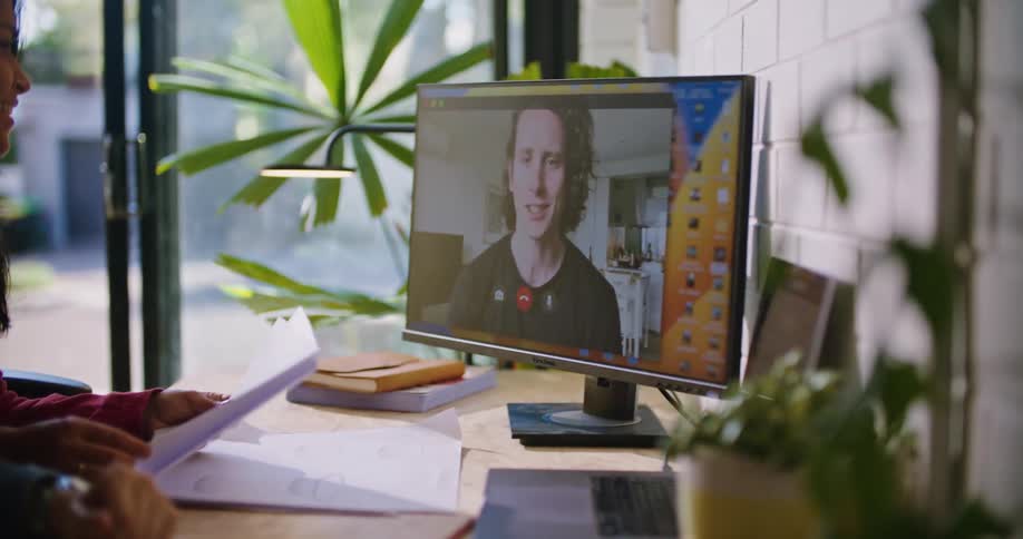 A man and a woman are sitting at a desk in front of a computer monitor as they participate in a video conference and she is holding papers in her hands