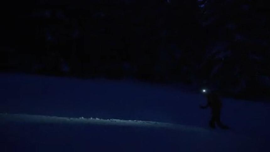 A mountaineer uses trekking poles to walk up a gentle slope covered in deep snow at night