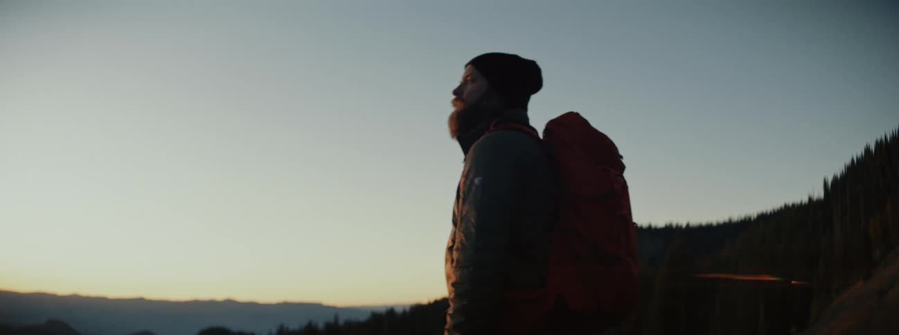 A man wearing a backpack is standing outside watching the sun set over the mountains
