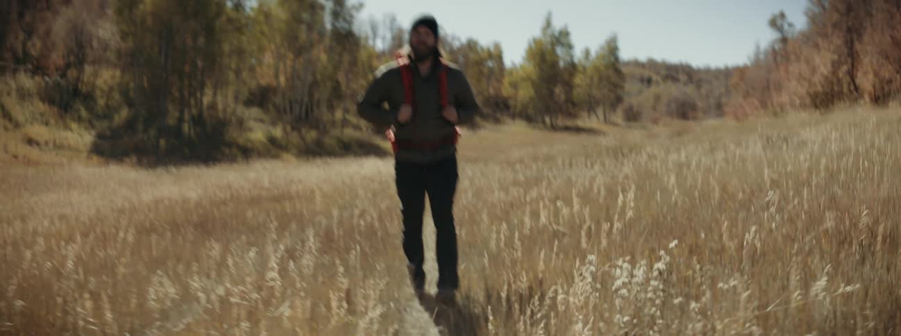 A man hikes wearing a backpack and hikes through an open field with trees surrounding it