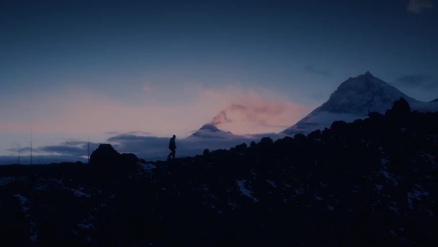 Man walking uphill is silhouetted against the snowy mountains and sky at dusk