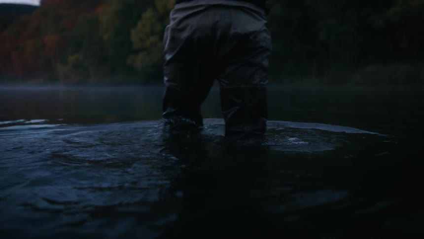 A man wearing tall fishing boots is walking through the water and there are trees with leaves in fall colors along the shore