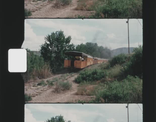 A passenger train with open cars travels through a rural area under partly cloudy skies