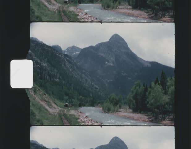 Old film footage of train tracks next to a river with a mountain range in the background