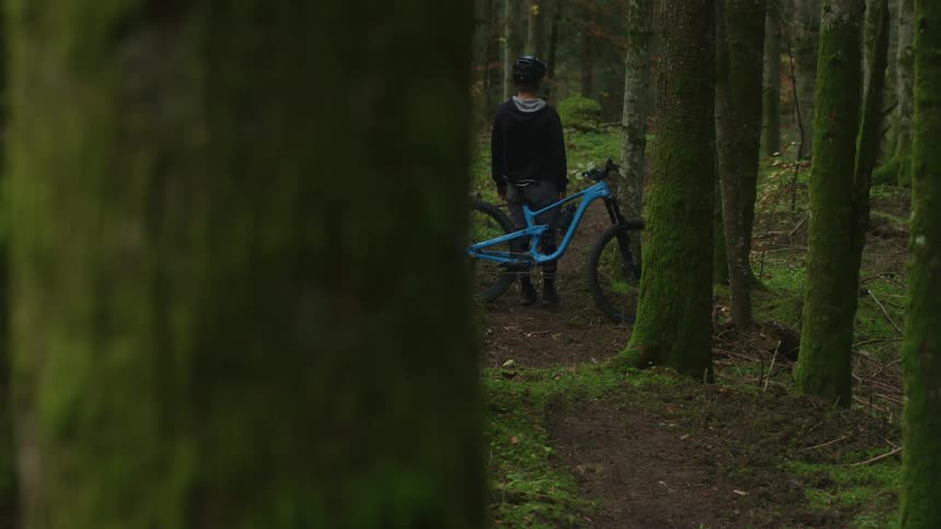 A teenage boy is standing next to his bicycle on a trail in the woods and he picks up the bike and turns it in another direction
