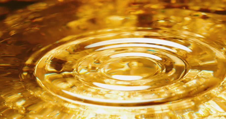 A clear sphere falls into a golden liquid and creates a rippling ring on the surface and splashes back up