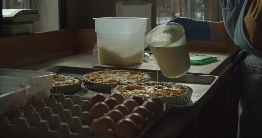 Someone pouring a milky substance from a jug onto a pizza while two other pizzas are visible on the table the scene also includes eggs in a crate and white bowls on the table