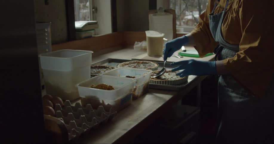 A woman is standing over a table holding a spoon as she fills pie shells for pumpkin pie
