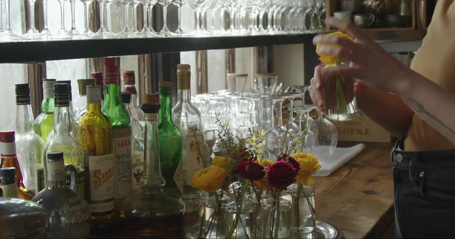 Female bartender stands near counter strewn with liquor and adjusts flowers in water sitting on a tray