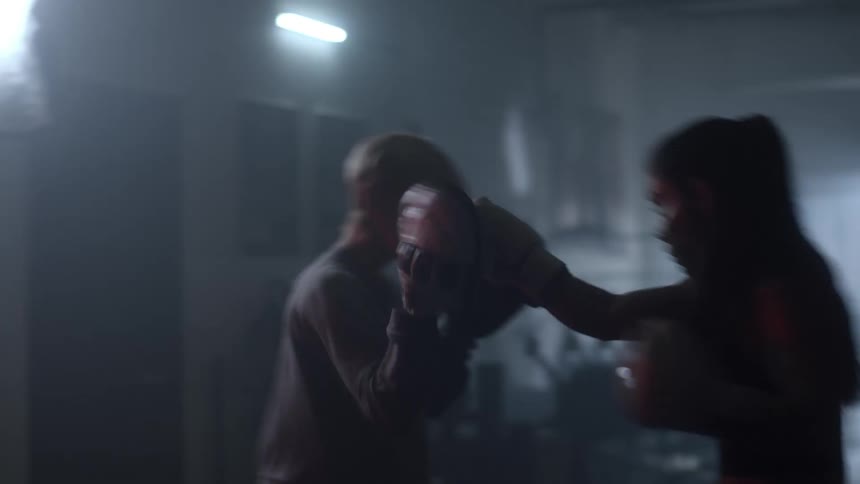 A woman training in a dark gym punching pads with a partner