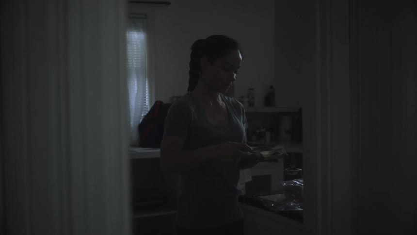 A woman is standing in the kitchen holding a knife as she spreads peanut butter on a slice of bread