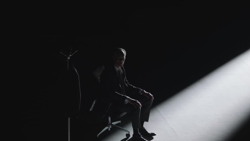 A man wearing a suit sitting in an office chair slapping himself in the face stands and walks away