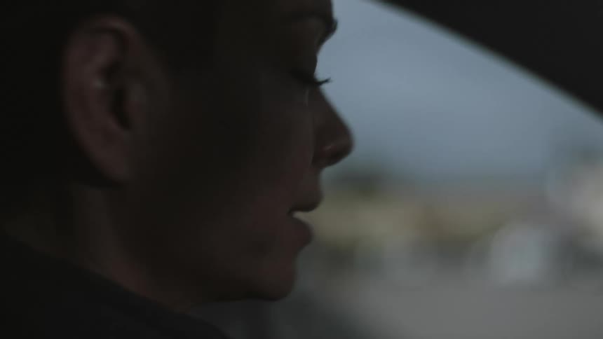 A woman with shaved head and pony tail sits in a parked car in the drivers seat and takes a deep breath as for preparation and gets out of the car