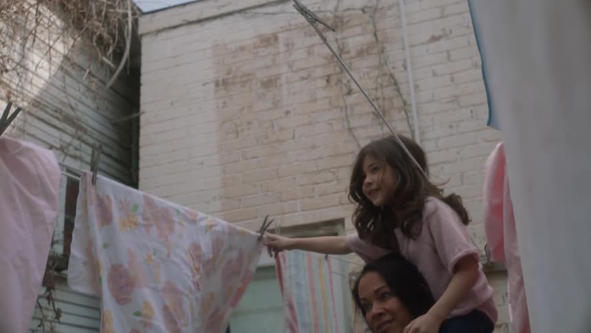 Daughter sitting on mothers shoulders as they walk in between clotheslines with linens hanging to dry