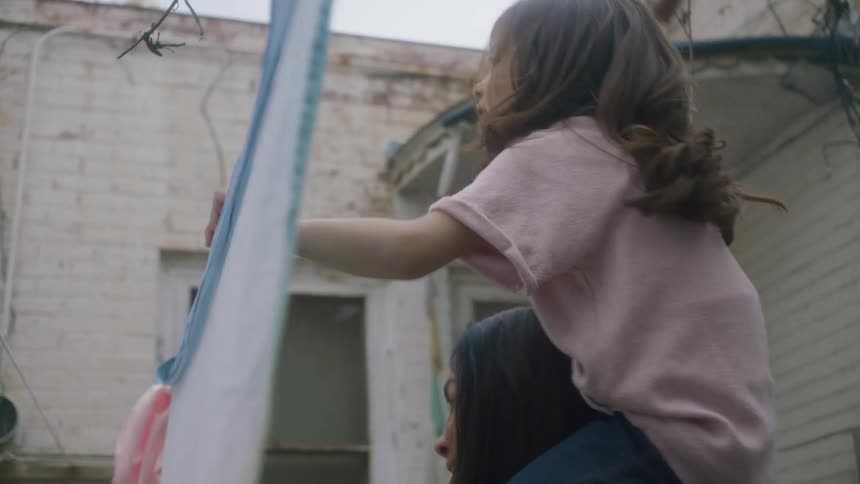 A little girl is riding on her mothers shoulders as they hang sheets on the clothes line