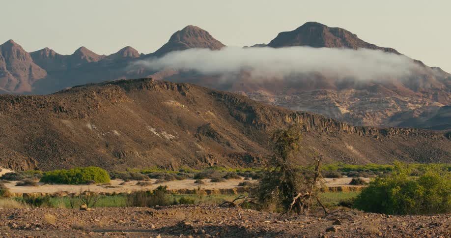 A dirt plain next to a hill with mountains in the background framed by clouds and trees in the foreground