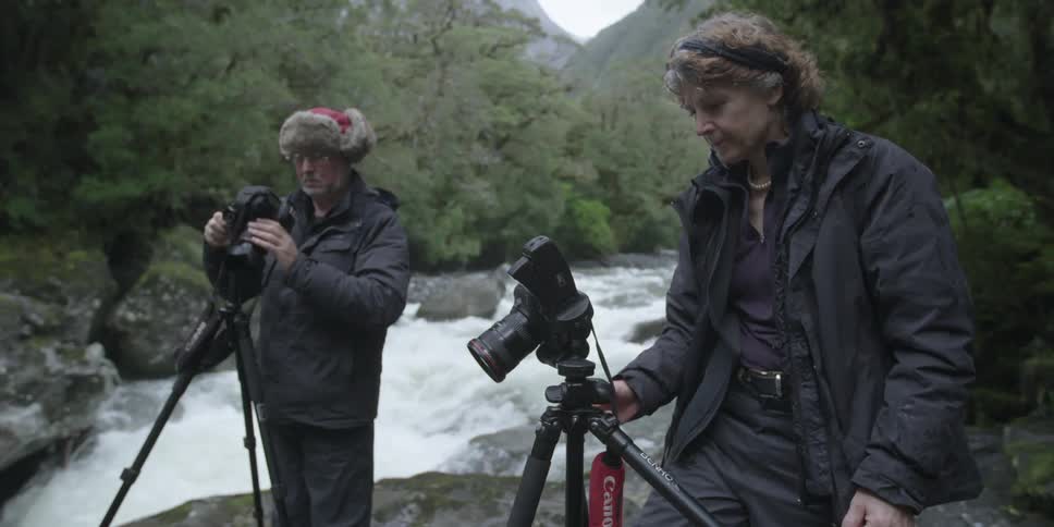 A man and woman adjust their cameras near a river