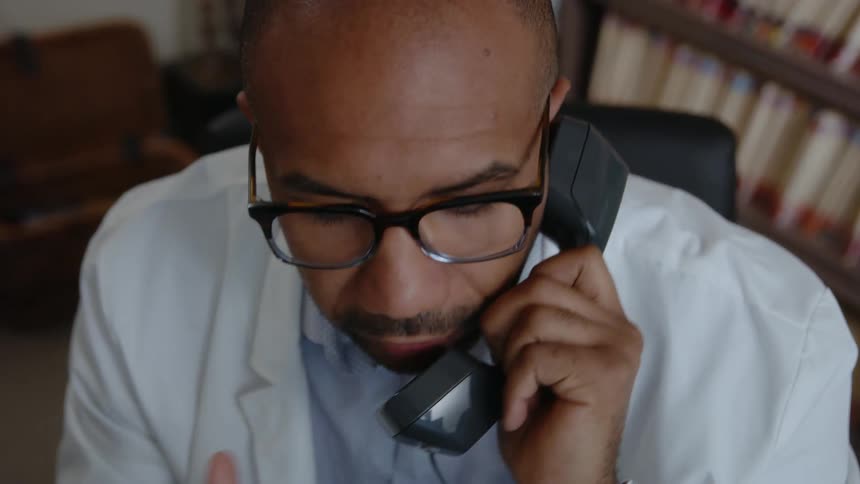 A man wearing a white lab coat holds a telephone receiver and smiles as he talks