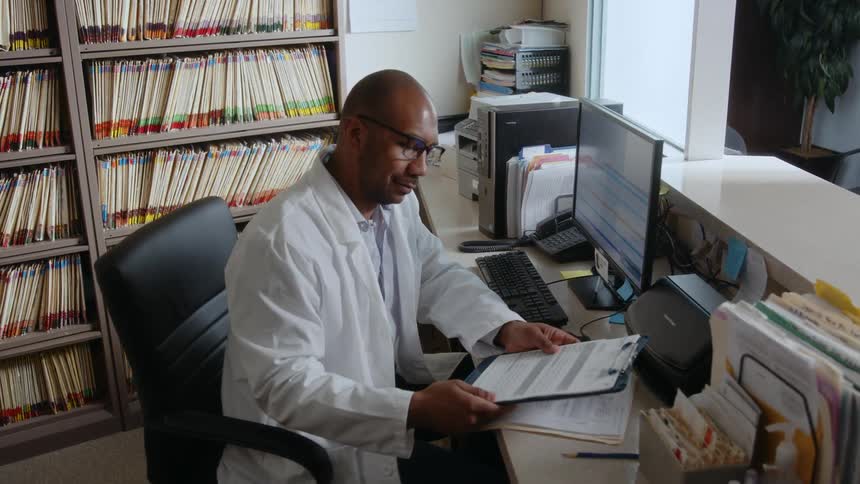 A black man in his office engaged in writing something into a book he then picks up the book to check the details and subsequently looks into the desktop computer placed on the table