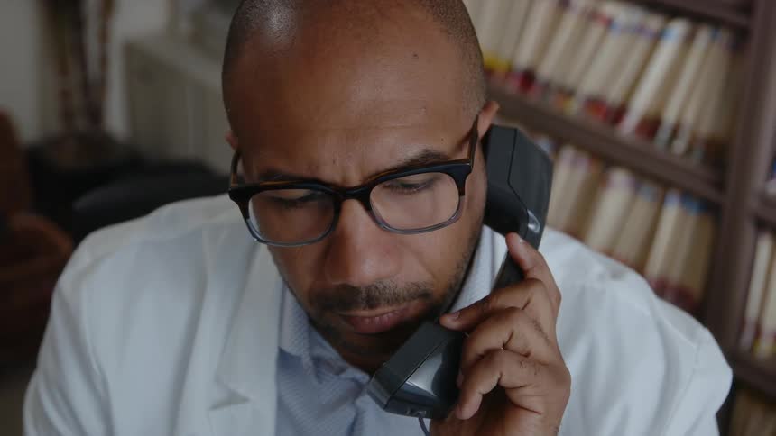 A man wearing a white lab coat is holding a telephone receiver and smiles as he speaks
