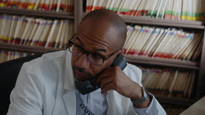 A man wearing a white lab coat is sitting at a desk as he holds a telephone receiver and touches his glasses with the other hand as he speaks