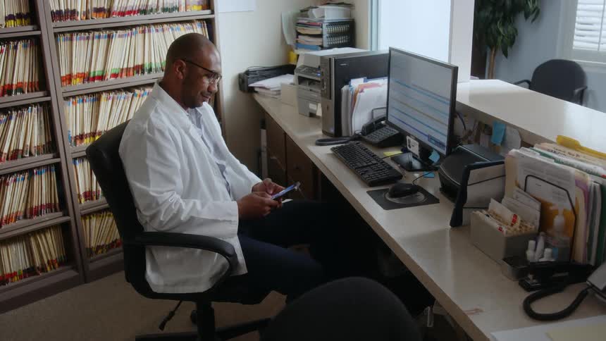 A male doctor sitting at his office desk using his cell phone in front of a computer screen