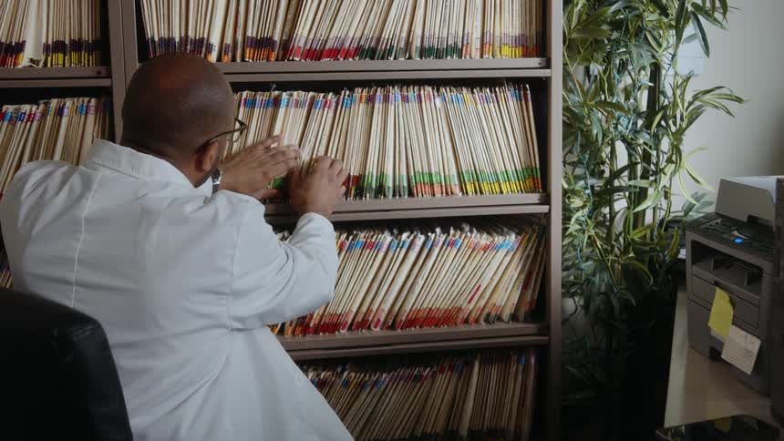 A man wearing a white lab coat is sitting at a desk working on a computer as he turns and finds a folder on the shelf behind him