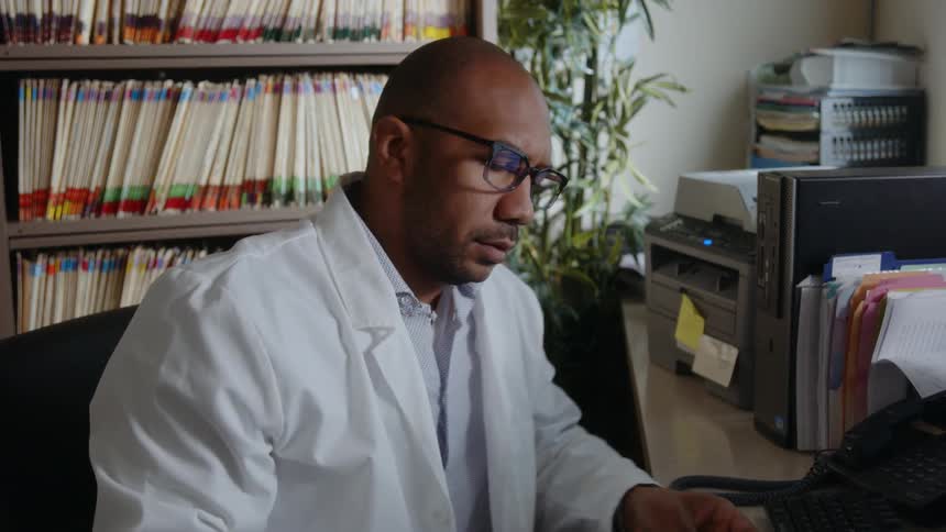A male doctor sitting in an office chair grabbing a paper off the printer and looking at it