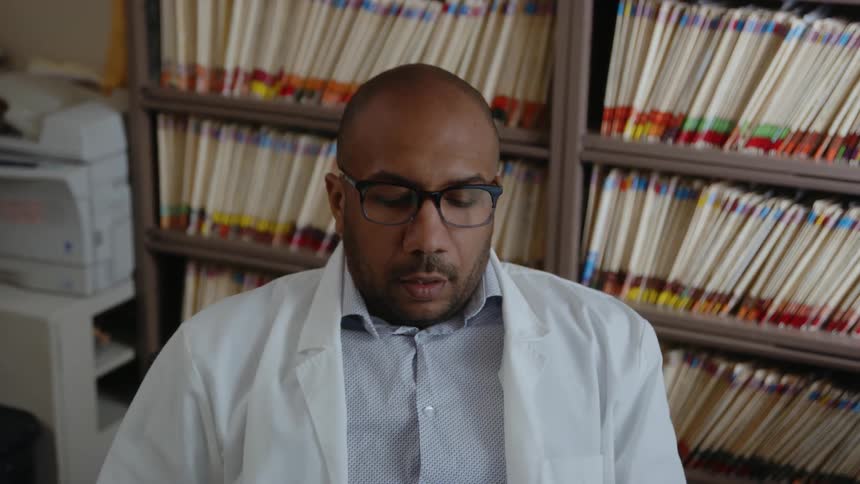 A man wearing a white lab coat sits down in the chair in his office and turns his head as he thinks