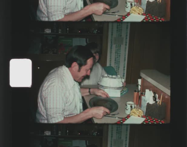 A father and son sitting at a counter the boy with a cake in front of him while holding up a paper with 53 on it