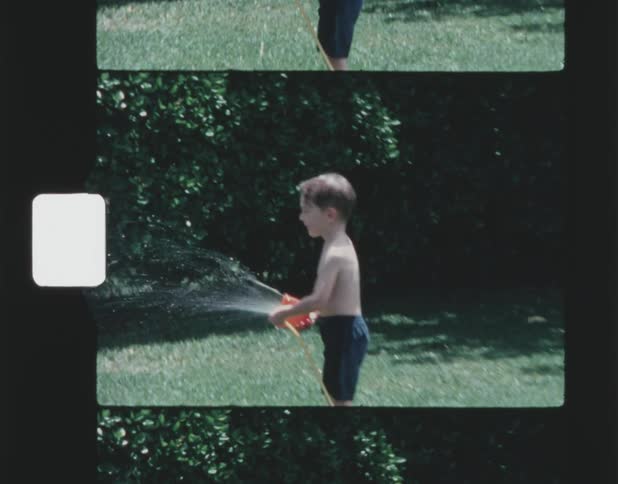 A little boy is outside in a bathing suit holding a sprinkler and spraying water in front of him