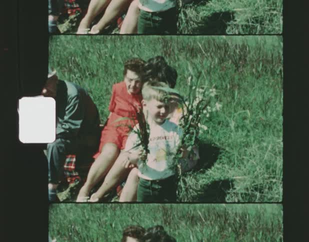 A young woman sitting next to her elderly parents in the grass while a young boy brings flowers over