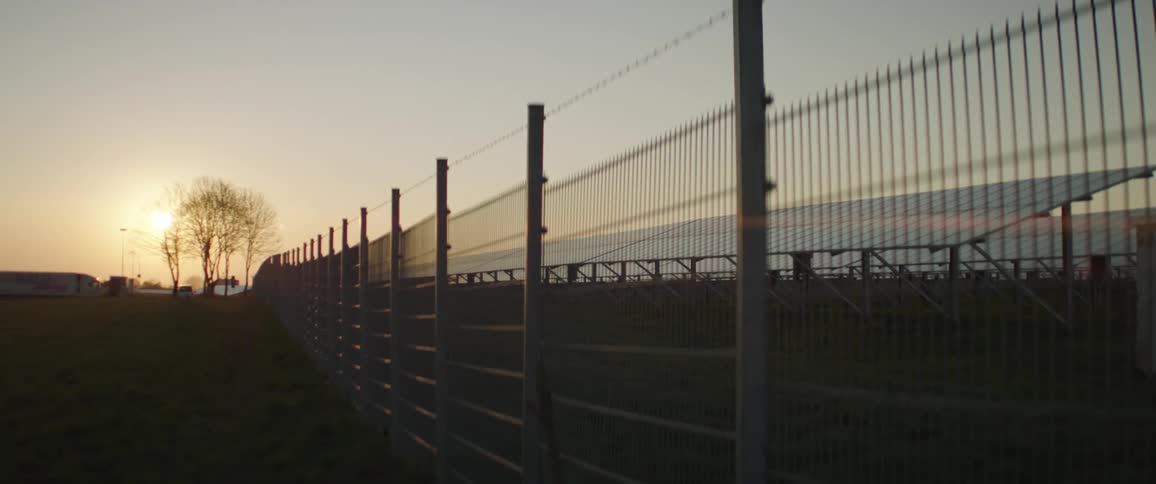 A fence is focused on with a semi truck on the far background at dusk or dawn