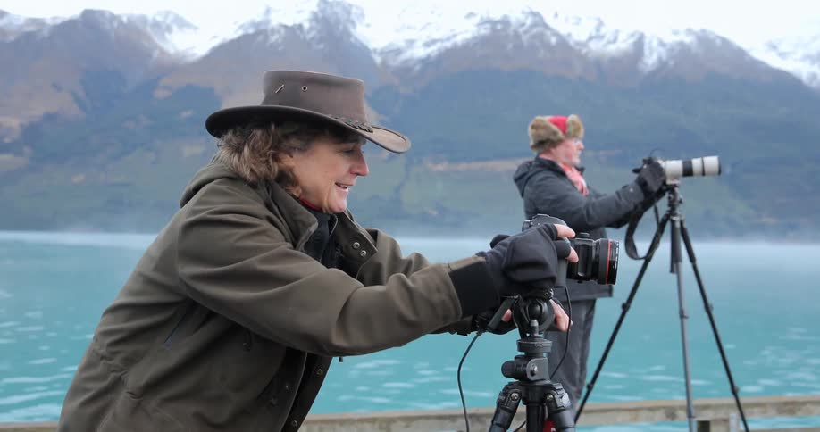Two people are using cameras on a pier near a mountain range