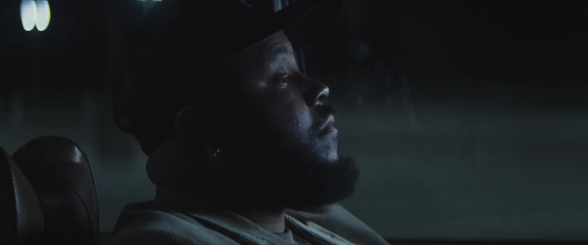 A man sitting inside a convertible car at night looks ahead and then to the side pensive