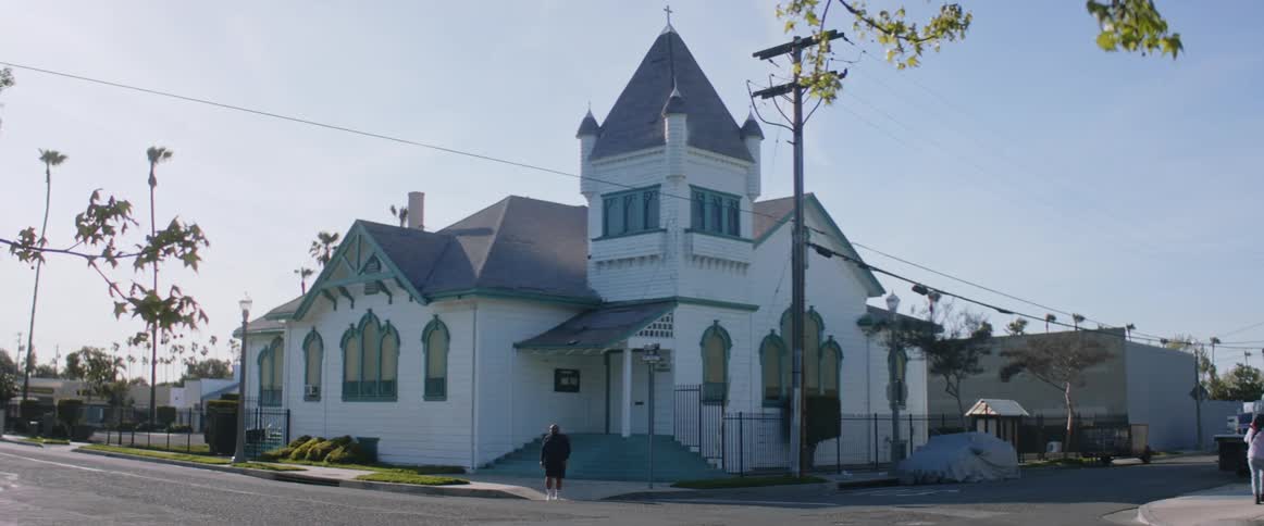 A large man wearing a black sweatshirt walks across an intersection next to a historic church